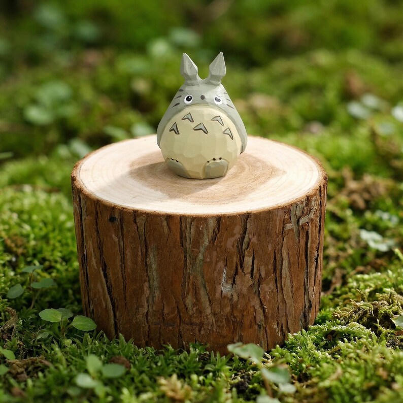 May include: A gray and white Totoro figurine with a light yellow belly, perched on a small, round wooden stump. The stump is surrounded by green moss. The figurine has pointed ears and distinctive markings.