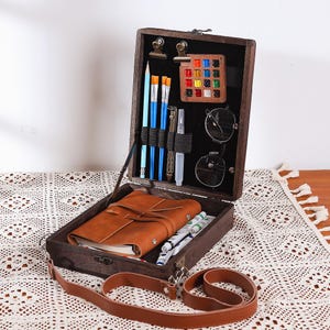May include: A dark wooden artist's box, open to reveal art supplies. Inside are paintbrushes, pencils, a palette of colours, glasses, a leather-bound journal, and tubes of paint. A brown leather strap is attached to the box.