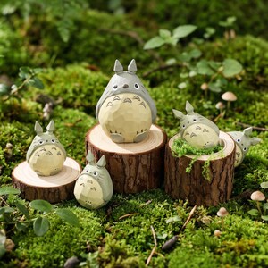 May include: A collection of gray and cream-colored Totoro figurines, each with a unique pose, displayed on small wooden platforms. The figurines are set amidst a bed of green moss and small mushrooms, creating a miniature forest scene.