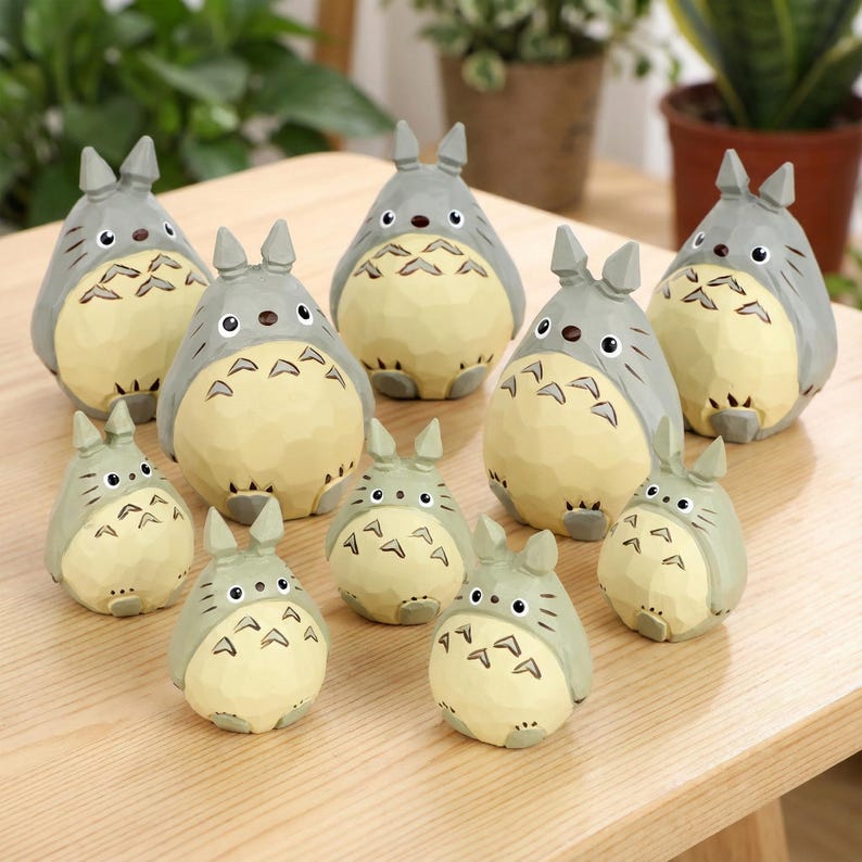 May include: A collection of gray and cream-colored Totoro figurines of varying sizes. The figurines have a textured appearance and feature the iconic Totoro design with pointed ears and a round belly. They are arranged on a light-colored wooden surface.