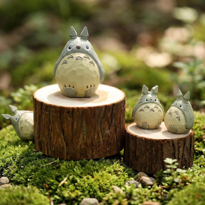 May include: Wooden figurines of the character Totoro from the animated film, My Neighbor Totoro. The figurines are light gray and beige, and are displayed on small wooden tree stumps. The background is a natural setting with green moss.