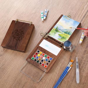 Op de afbeelding: Een houten aquarelverfset met een scharnierend deksel, een palet en een selectie van kleurrijke verven. De set bevat een schilderij, penselen en verftubes. Het deksel is gegraveerd met een decoratief ontwerp en de letter "M".