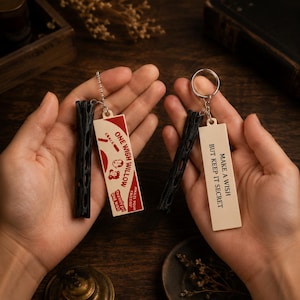 May include: Two keychains featuring a black cylindrical textured piece and a rectangular tag. One tag is cream with red accents and the text ONE WISH WILLOW, while the other is cream with black text reading MAKE A WISH BUT KEEP IT SECRET.