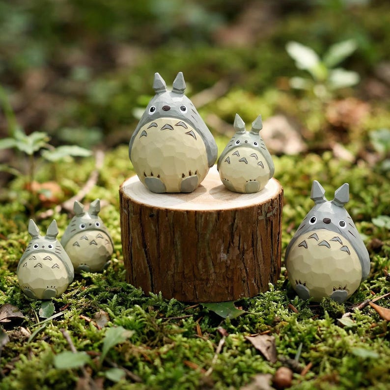 May include: A collection of five Totoro figurines in various sizes. The figures are gray and cream-colored, with distinctive ear shapes and facial features. They are arranged on a wooden stump and in a bed of green moss.