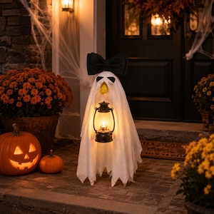 May include: A white ghost decoration featuring a large black bow on top, black eye cutouts, and a small yellow beak. The ghost figure holds a black metal lantern with a glowing warm light, standing on a stone porch with pumpkins and flowers.