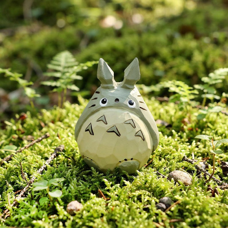 May include: A light green Totoro figurine with black markings and a white belly, set against a backdrop of green moss and foliage. The figurine has pointed ears and a series of arrow-shaped markings on its chest.