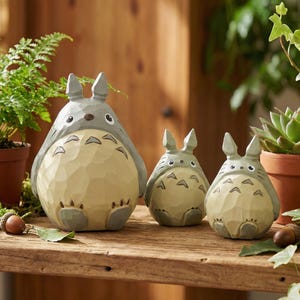 May include: Three carved wooden Totoro figurines in varying sizes. The largest is gray and beige, with a dark gray nose and eyes. The smaller figures are light green and beige. The figurines are on a wooden surface.