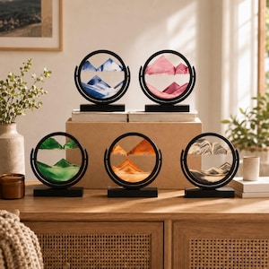 May include: Five circular sand art displays with black frames and rectangular bases. Each piece contains colored sand forming mountain-like landscapes in blue, pink, green, orange, and black and white, encased between two clear glass panes.