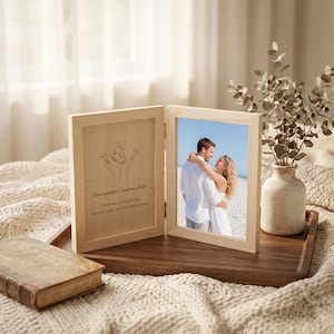 May include: A light-coloured wooden double photo frame featuring a hinged design. One side displays a line art illustration of hands holding flowers with the text Thoughtful Custom Gift, while the other side holds a photograph of a couple on a beach.