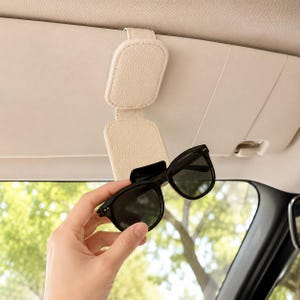 May include: A beige, textured faux leather car visor sunglasses holder with a magnetic closure flap. The holder is attached to a vehicle sun visor and is currently holding a pair of black-framed sunglasses with dark, tinted lenses.