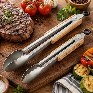 May include: Two stainless steel kitchen tongs with light wood handle grips and black circular hanging loops. One pair of tongs features the engraved text "Bestes Papa & Grillmeister" and the other pair features the engraved text "Mike's Grill 2026".