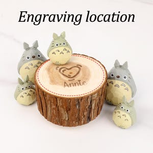 May include: A wooden ring holder with a heart and the name "Annie" engraved on it, surrounded by several small, light green and gray Totoro figurines. The text "Engraving location" is at the top.