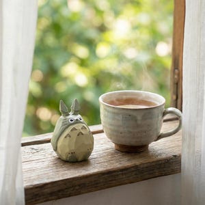 May include: A wooden figurine of a Totoro character sits next to a ceramic mug filled with tea. The mug is light blue and has a brown base. Steam rises from the tea, and the scene is set on a wooden windowsill with a blurred green background.