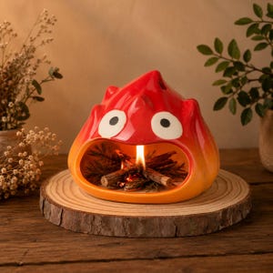 May include: A glossy red and orange ceramic flame-shaped sculpture with two large white eyes and a wide open mouth. Inside the hollow opening, a small flame burns among several thin wooden twigs, creating a miniature campfire effect.