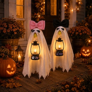 Pode incluir: Duas decorações de fantasmas brancos com bordas inferiores serrilhadas, cada uma segurando uma lanterna preta com uma luz brilhante. Um fantasma tem um grande laço rosa e o nome Lily, o outro tem um laço preto e o nome Emma na lanterna.