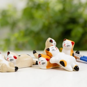 Op de afbeelding: Een verzameling keramische kattenbeeldjes in verschillende poses. De katten hebben witte, oranje en bruine vacht met zwarte markeringen en rode accenten. De beeldjes zijn klein en lijken decoratieve items te zijn.