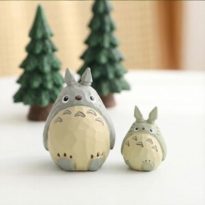 May include: Two gray and cream-colored Totoro figurines, one large and one small, stand in front of two miniature green pine trees. The figurines have distinctive features, including large eyes and a belly pattern. The scene evokes a whimsical, forest-like setting.