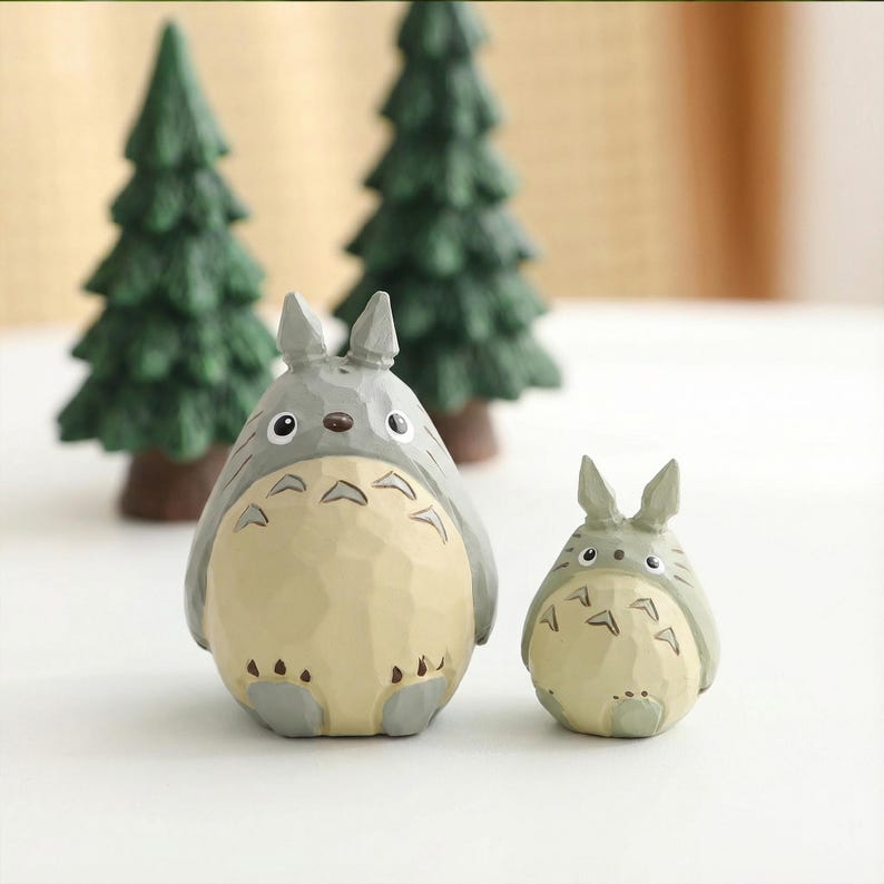 May include: Two gray and cream-colored Totoro figurines, one large and one small, stand in front of two miniature green pine trees. The figurines have distinctive features, including large eyes and a belly pattern. The scene evokes a whimsical, forest-like setting.