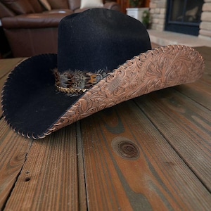 Puede incluir: Un sombrero de vaquero negro con un borde decorativo de color cobre con un patrón floral en relieve. El sombrero tiene una pluma y un detalle de banda dorada. El sombrero está sobre una superficie de madera.