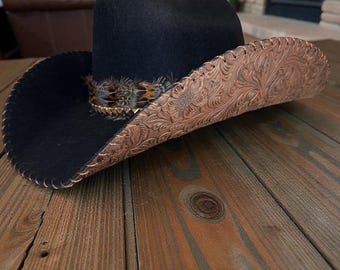 May include: A black cowboy hat featuring a wide, upturned brim with intricate floral tooling in a tan leather finish. The hat is accented with a band of multi-colored feathers and a metallic chain detail encircling the crown.