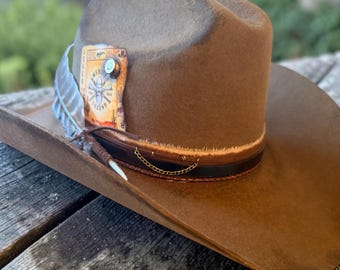 May include: A brown felt cowboy hat featuring a leather band with a thin gold chain, a white feather, and a rectangular patch on the side. The patch displays the text American Legend, a graphic of crossed rifles, and a small metallic bullet casing accent.