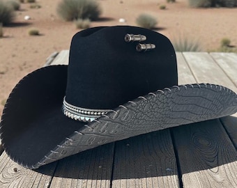 May include: A black cowboy hat with a decorative band and silver accents. The hat has a textured brim and two silver embellishments on the crown. The hat is sitting on a wooden surface.