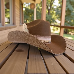 Könnte beinhalten: Ein brauner Cowboyhut mit Schlangenmuster-Rand und Federakzent. Der Hut hat ein hellrosa Band mit goldfarbenen Nieten und einen genähten Rand. Der Hut liegt auf einer Holzoberfläche.