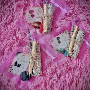 Może przedstawiać: Trzy przezroczyste plastikowe woreczki z zestawami rytualnymi. Każdy zestaw zawiera pęczek szałwii, patyczek palo santo, kryształ, małą świeczkę i gałązkę lawendy, związane sznurkiem. Dołączono karty z napisami CONFIDENCE, PROSPERITY oraz PROTECTION.