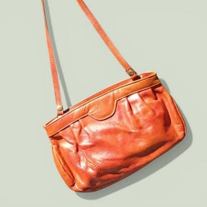 Puede incluir: Bolso de hombro vintage de cuero naranja con una correa larga. El bolso tiene forma rectangular con la parte superior curvada y un cuerpo texturizado y fruncido. La correa es fina y ajustable, y el bolso parece estar en buen estado.