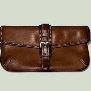 Puede incluir: Bolso de mano de cuero marrón con hebilla plateada. El bolso tiene costuras blancas y una correa de muñeca. El cuero tiene un ligero brillo.