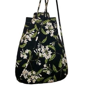 Puede incluir: Bolso tote negro con un estampado floral de flores blancas y hojas verdes. El bolso tiene dos asas y una correa larga para llevar al hombro o cruzado. El bolso está hecho de una tela suave y acolchada.