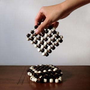 Puede incluir: Un posavasos cuadrado hecho de cuentas de madera marrones y blancas alternas. Un posavasos se levanta, mientras que otro descansa sobre una superficie de madera. Las cuentas están dispuestas en un patrón de cuadrícula, creando una superficie texturizada. Para la decoración del hogar.