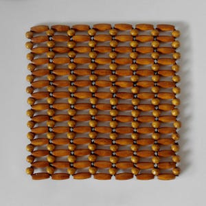 May include: A square trivet constructed from a grid of light brown wooden beads and elongated wooden cylinders. The components are connected by dark strings, creating a flexible, open-weave pattern with a natural wood grain finish throughout.