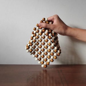 Puede incluir: Un objeto cuadrado con cuentas compuesto por cuentas de madera claras y oscuras alternas. Las cuentas están dispuestas en un patrón de cuadrícula, creando una superficie texturizada. El objeto está sostenido por una mano contra un fondo blanco y una superficie marrón.