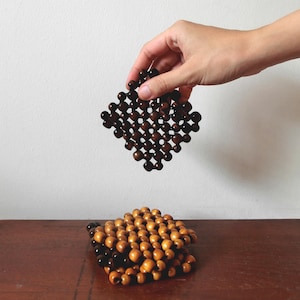以下が含まれることがあります： ダークブラウンの木製ビーズを格子状に並べて作られた正方形のコースター。その下には、ライトタンとダークブラウンの木製ビーズを組み合わせた同様のコースターが積み重ねられており、同じ格子状の構造とビーズのデザインが特徴です。