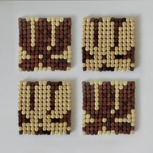 Puede incluir: Cuatro posavasos cuadrados construidos con pequeñas cuentas de madera individuales dispuestas en un patrón de cuadrícula. Cada posavasos presenta un diseño geométrico creado alternando cuentas de color beige claro y marrón oscuro.