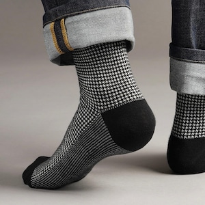 May include: A pair of black and white houndstooth socks with a black heel and toe. The socks are paired with rolled-up denim jeans. The socks have a striped pattern on the lower part of the foot.