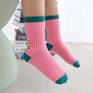 May include: Pink ribbed socks with green toes and cuffs. The socks have yellow smiley face designs on the side. The socks are designed for children.