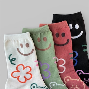 May include: Four pairs of crew socks in white, sage green, dusty rose, and black. Each sock features a repeating pattern of a smiling face, a simple flower outline, and a small squiggle. The designs are rendered in contrasting colours.