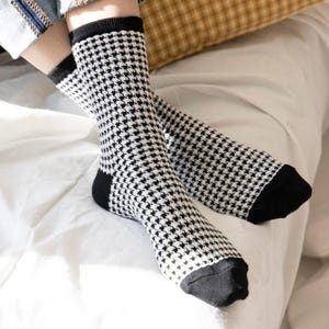 May include: A pair of mid-calf socks featuring a black and white houndstooth pattern. The socks have solid black cuffs at the top and solid black toe sections. The knit fabric displays a consistent repeating checkered design throughout the main body.