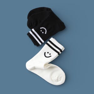 May include: Two pairs of socks, one black and one white, with a smiley face design. Each sock has two white stripes at the top. The socks are displayed on a blue background.
