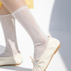 May include: Cream-colored knee-high socks with a delicate openwork design. The socks are paired with off-white ballet flats. The socks have a ribbed cuff at the top and are slightly scrunched at the ankles.