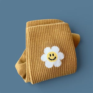 May include: Mustard yellow ribbed knit leggings with a white daisy applique featuring a yellow smiley face. The leggings are folded, showcasing the texture and design. The background is a solid blue.