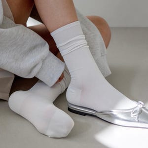 Puede incluir: Calcetines blancos con puño acanalado, combinados con bailarinas plateadas. Los calcetines están ligeramente arrugados. Los zapatos tienen un acabado metálico plateado y un pequeño lazo.