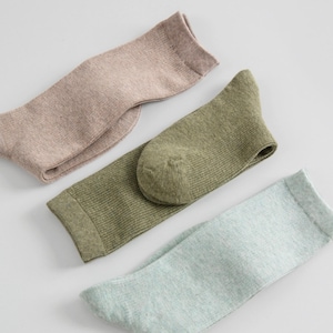 May include: Three pairs of socks in neutral colors are displayed. One pair is light brown, another is olive green, and the third is a light blue-green. The socks are made of a ribbed knit fabric and are folded neatly.