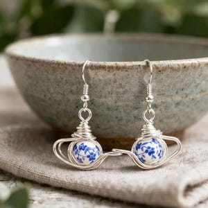 Può includere: Un paio di orecchini a forma di teiera argentati. Ogni orecchino presenta una piccola perla rotonda in ceramica bianca con motivi floreali blu, avvolta in filo metallico per formare una teiera. Gli orecchini pendono da ganci argentati.