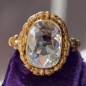 May include: A gold ring featuring a large, clear, cushion-cut gemstone. The ring has an ornate gold setting with floral and leaf detailing. The gemstone reflects light, creating a sparkling effect. The ring is displayed on a purple velvet surface.