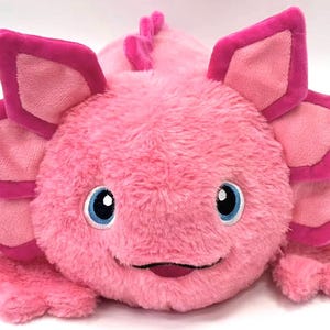 May include: A pink axolotl soft toy with petal-shaped gills edged in a darker pink. It has blue eyes, a smiling mouth, and small legs. The toy is designed to be soft and cuddly, a charming children's item.