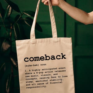 Puede incluir: Bolso tote beige con la palabra "comeback" impresa en negro. La definición de "comeback" está impresa debajo. El bolso es sostenido por una mano.
