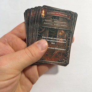 Könnte beinhalten: Ein Stapel dunkel getönter Spielkarten in einer Hand. Die Karten zeigen deutschen Text, darunter "LOCKRUF IM DUNKELN" und "PSYCHOLOGISCHE FALLE." Die Karten haben eine Vintage-Steampunk-Ästhetik mit Zahnrädern und einer Kerzenillustration.